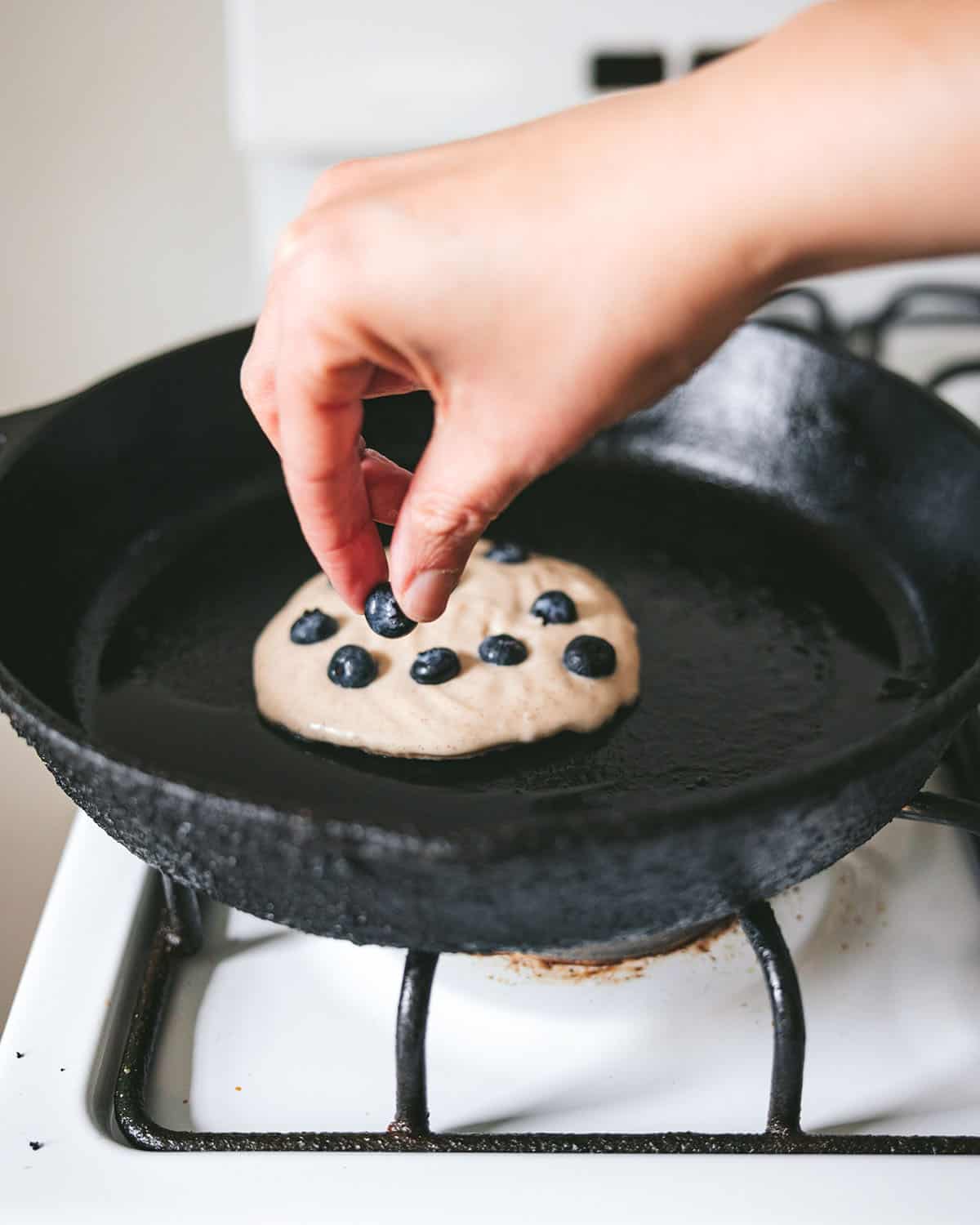 A hand placing blueberries into the raw top side of a cooking pancake on a cast iron skillet. 