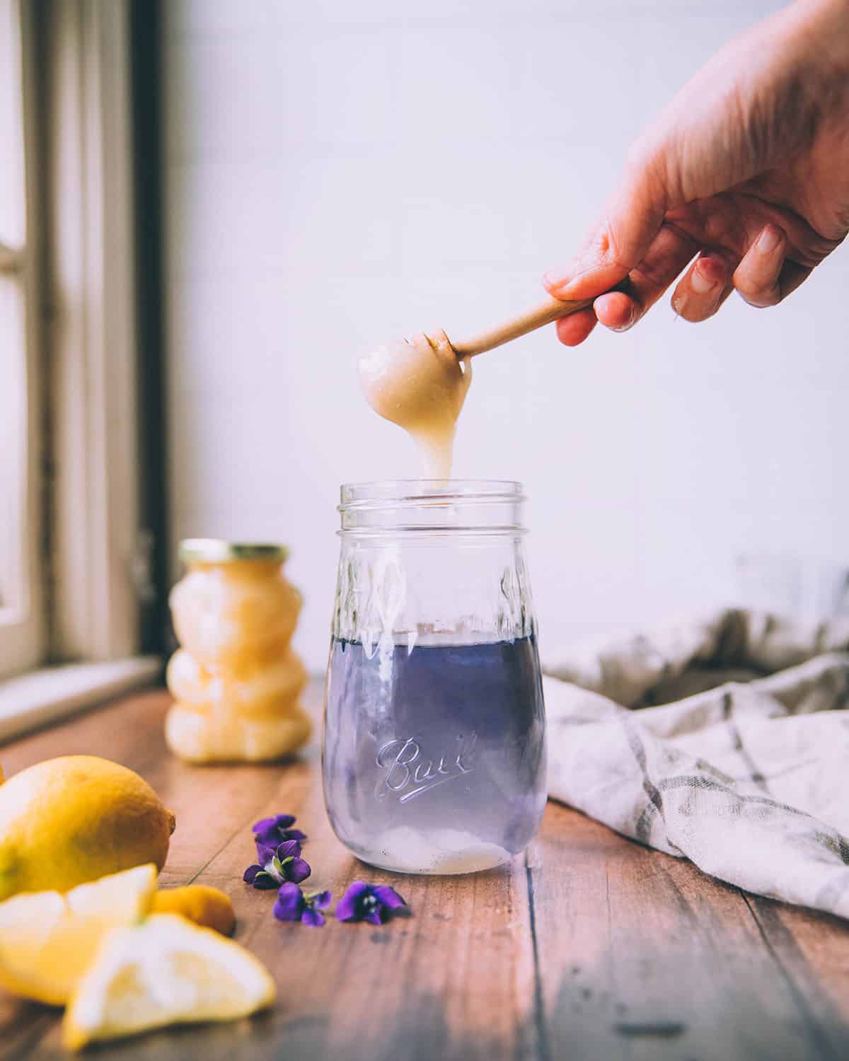 Honey drizzling into a jar of violet tea that is light purple color. On a dark wood surface with a honey bear, lemons, and wild violet flowers surrounding. 