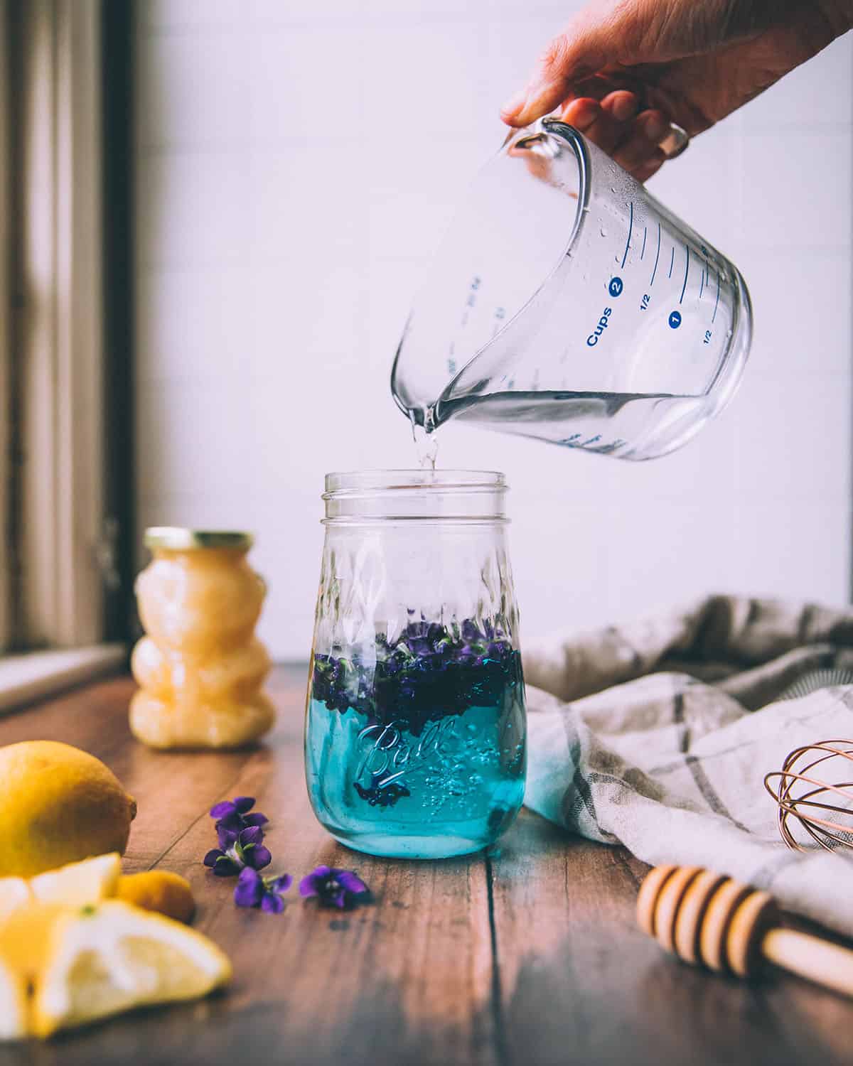 Hot water pouring over wild violet flowers in a jar, turning it a bright turquoise color. On a dark wood surface surrounded by a honey bear, honey stick, lemons, and wild violet flowers. 