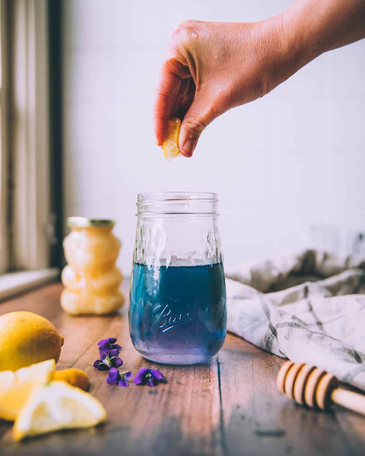 A jar of violet syrup changing color from purple at the bottom to turquoise at the top, with a lemon being squeezed into the jar. On a dark wood surface with a honey bear, honey wand, a lemon, and wild violet flowers surrounding. 
