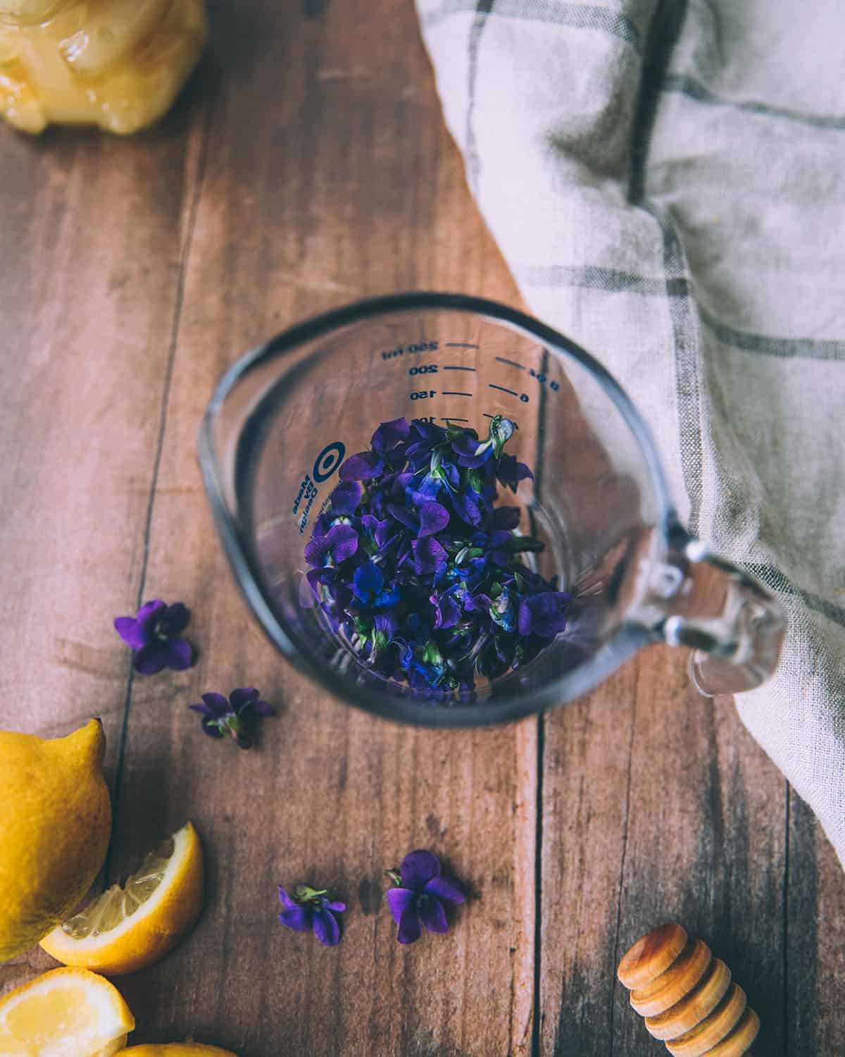 A glass pyrex measuring cup with a wild violet flowers in it, on a dark wood surface surrounded with violet flowers and a lemon. Top view.