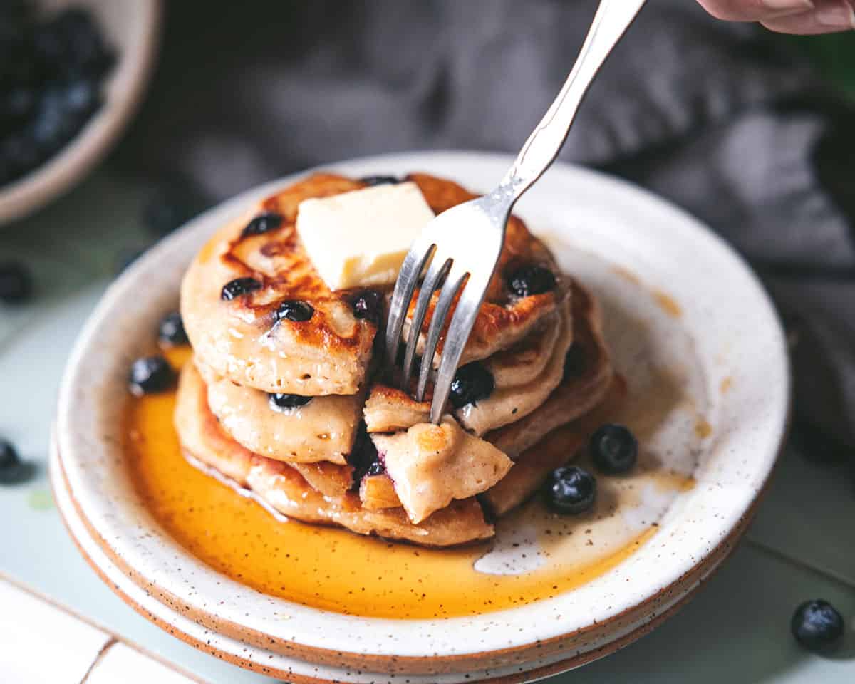 A plate of several sourdough pancakes with butter and syrup, with a fork cutting into them. 