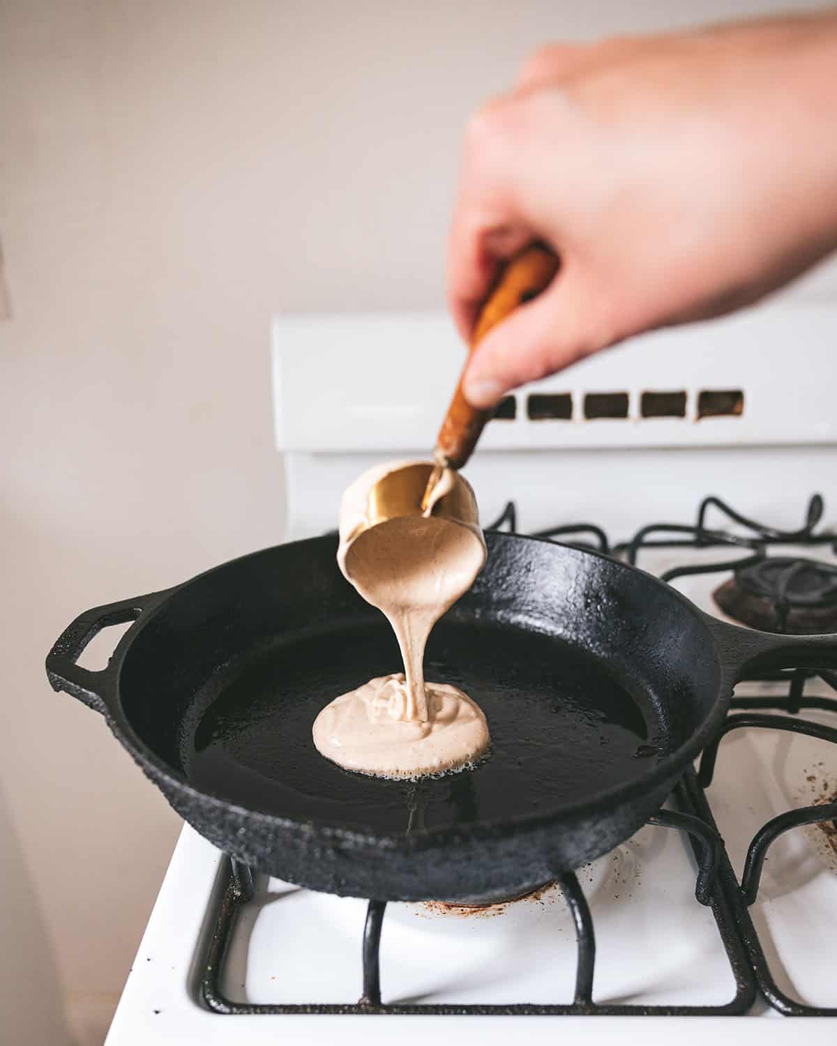 Pancake batter being poured with a measuring cup into a cast iron pan on a gas stove. 