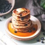 A stack of sourdough pancakes on a white plate with butter and syrup dripping down the sides, surrounded by blueberries and a bowl of blueberries in the background.