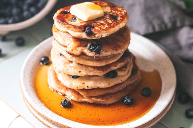 A stack of sourdough pancakes on a white plate with butter and syrup dripping down the sides, surrounded by blueberries and a bowl of blueberries in the background.