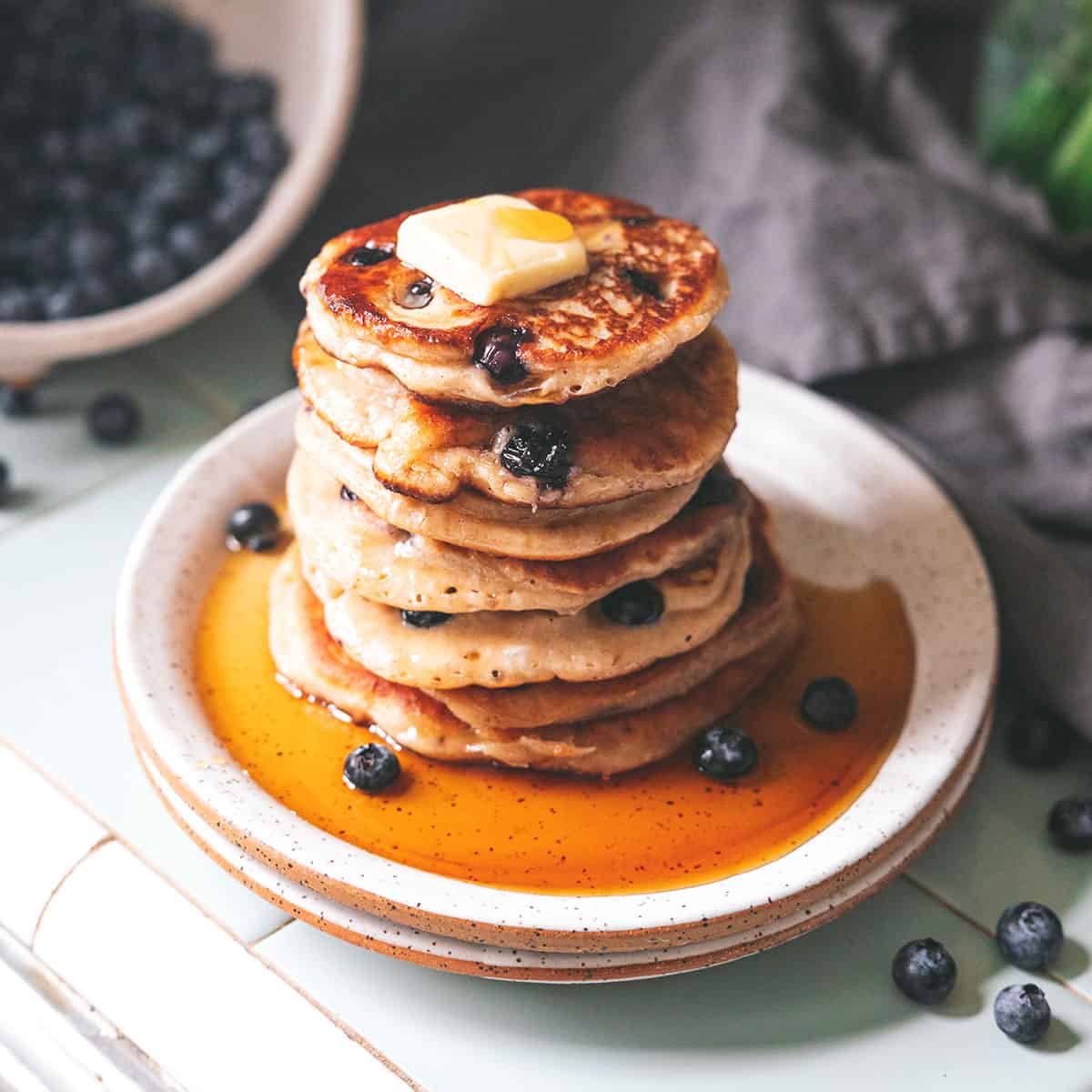 A stack of sourdough pancakes on a white plate with butter and syrup dripping down the sides, surrounded by blueberries and a bowl of blueberries in the background. 