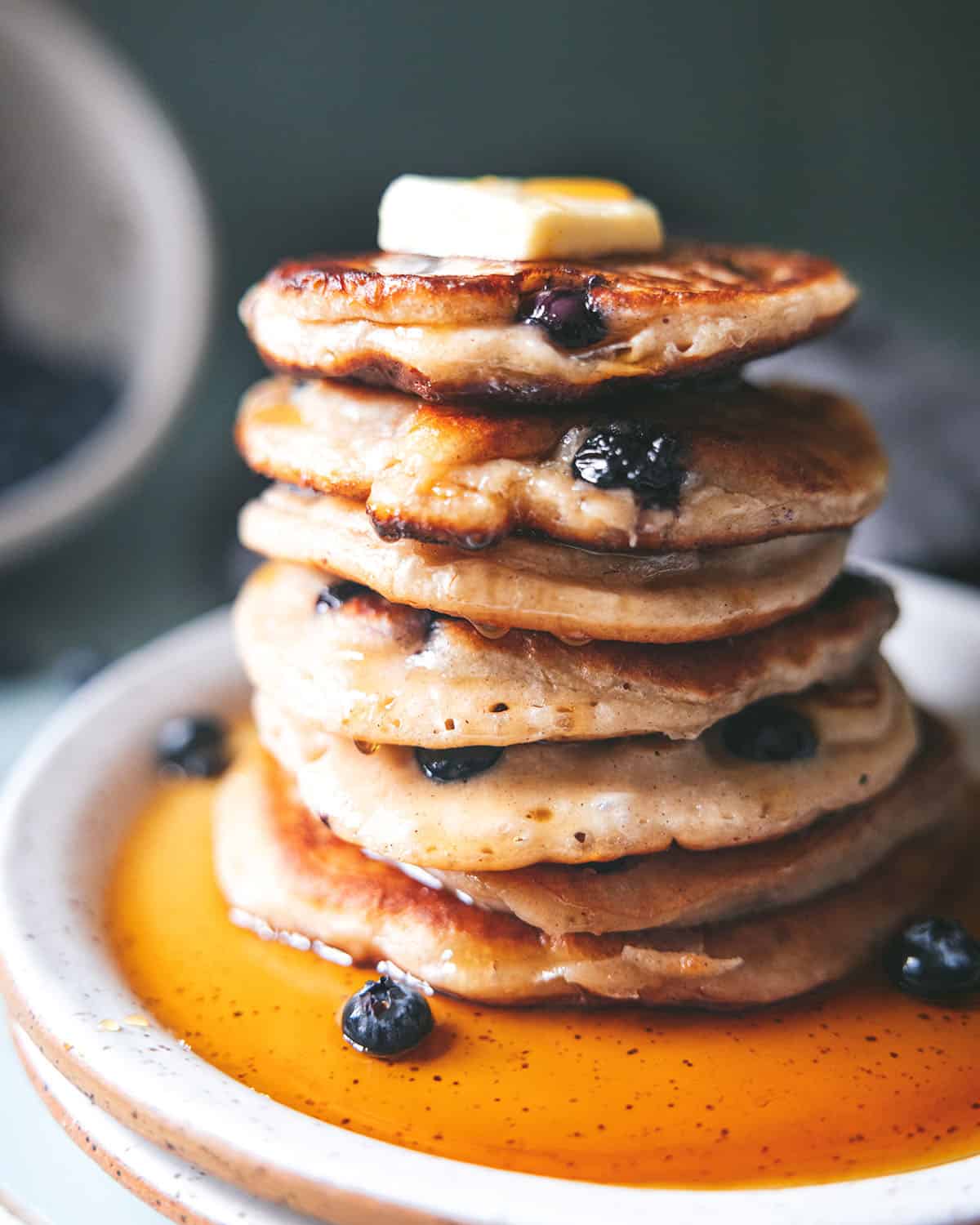 Close up side view stack of blueberry sourdough pancakes with butter on top and syrup drizzled on it and on the plate, with blueberries surrounding. 