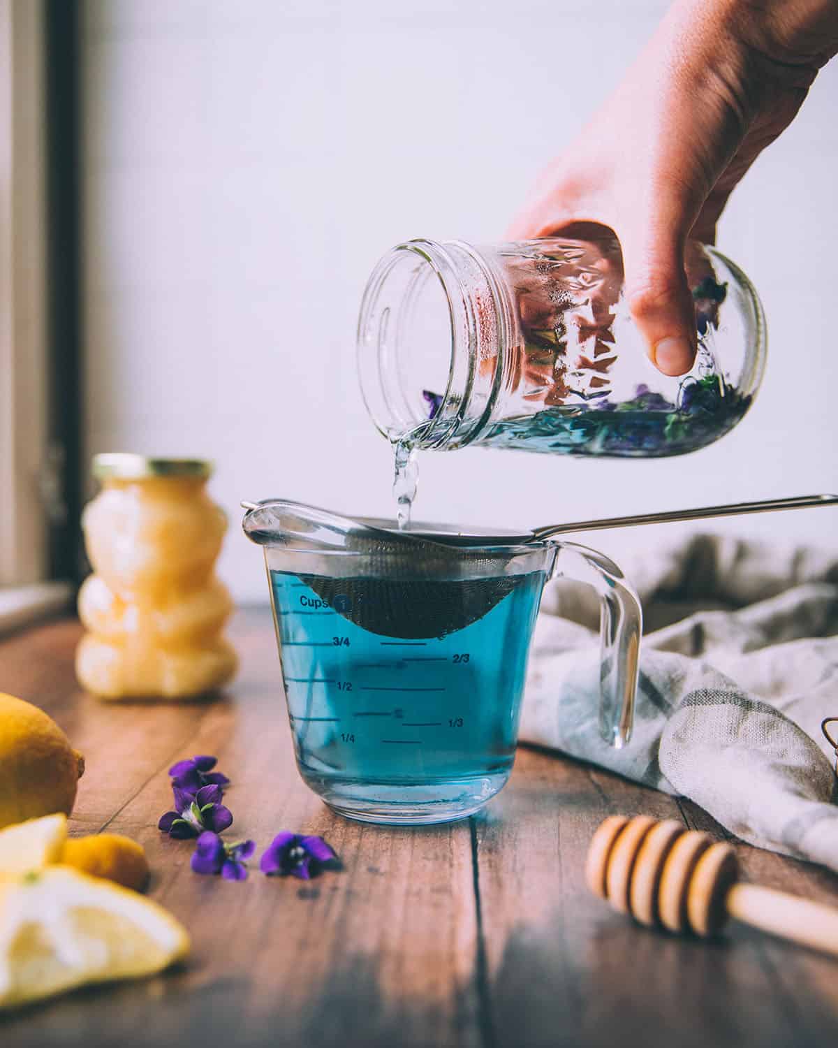 The mason jar of steeped violet flowers pouring over a glass measuring cup and a strainer to strain the flowers out. The liquid is turquoise. On a dark wood surface surrounded by a honey bear, a honey stick, lemons, and wild violet flower blooms. 