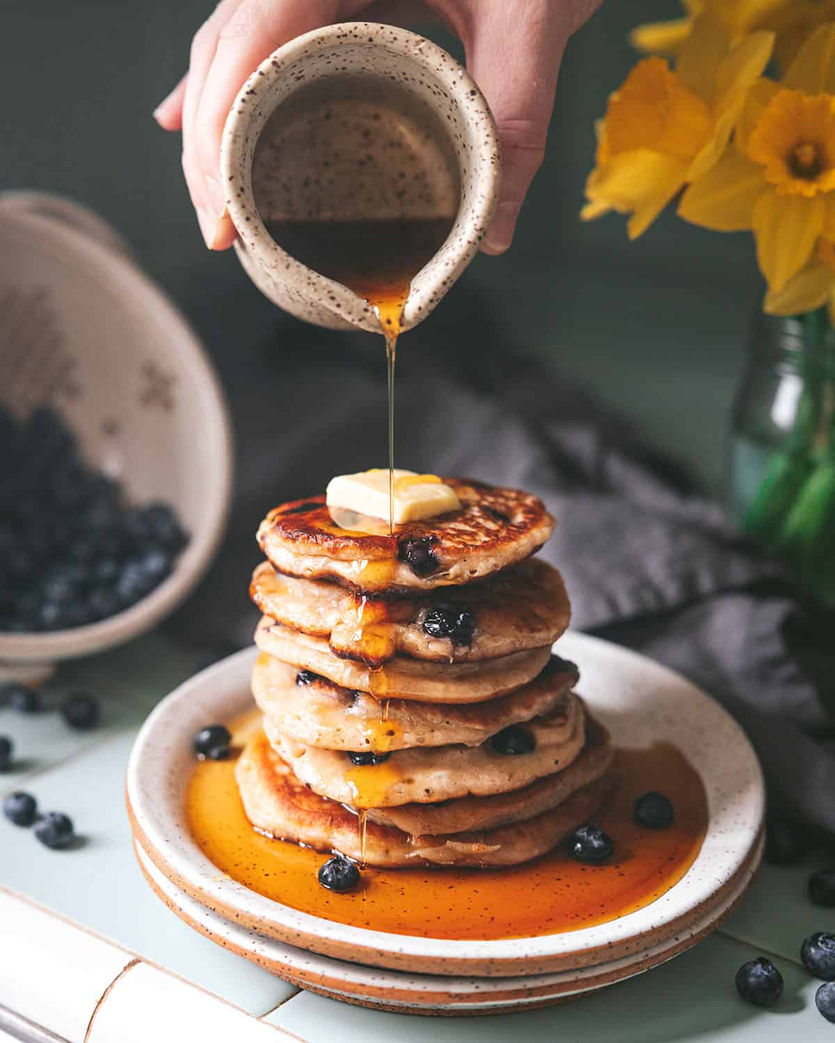 A tall stack of sourdough pancakes with a pat of butter, and syrup being drizzled on top of them. 