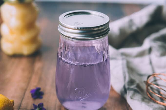A jar of wild violet syrup that is a light purple color, sitting on a wood surface with a honey bear in the background, and wild violet flowers surrounding.