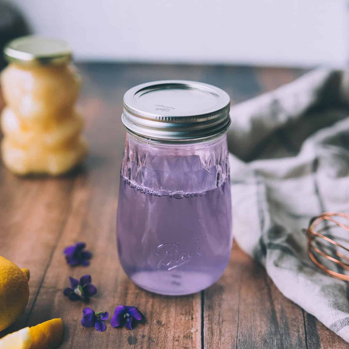 A jar of wild violet syrup that is a light purple color, sitting on a wood surface with a honey bear in the background, and wild violet flowers surrounding.