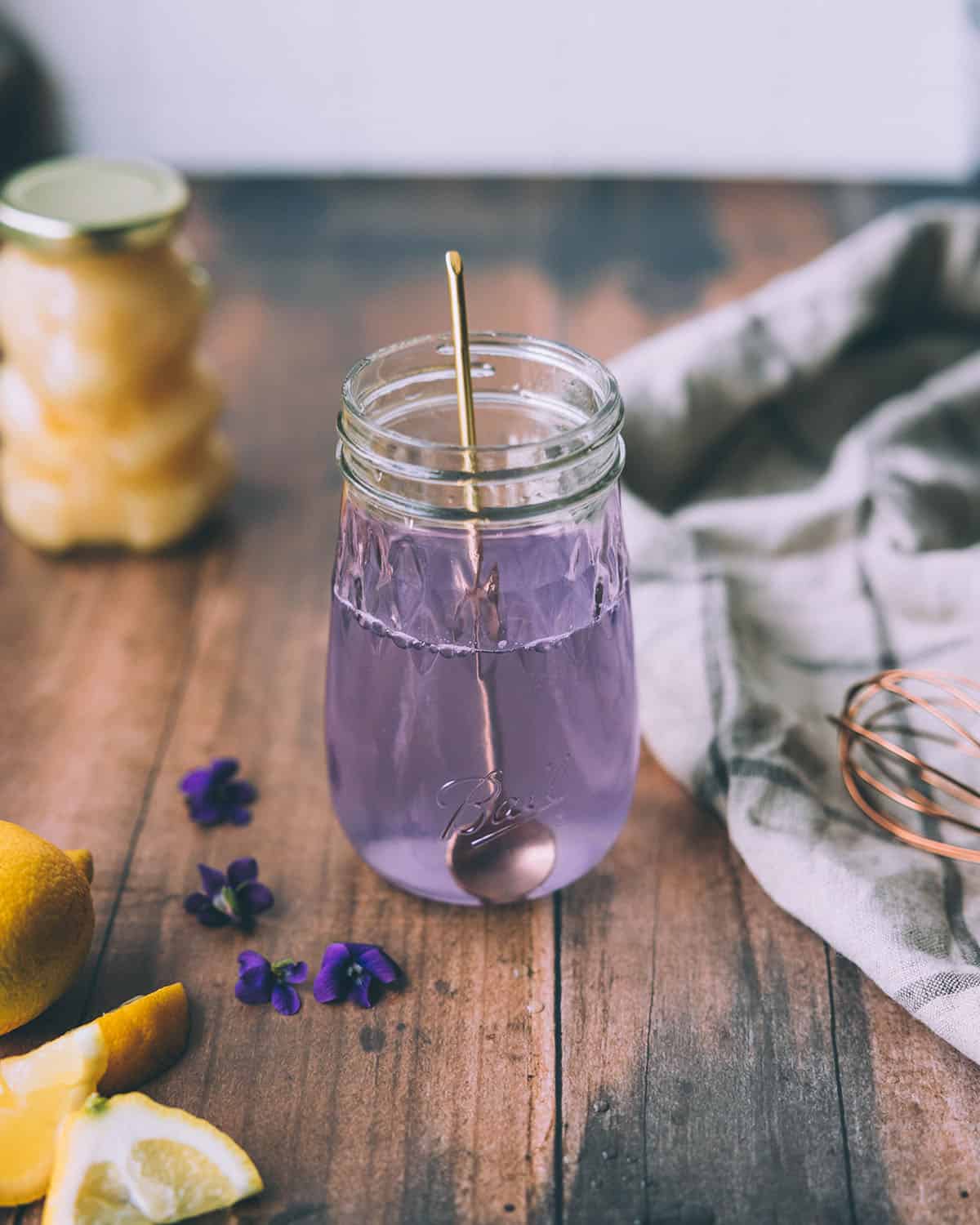 Wild violet syrup in a jar with a gold spoon, showing a light violet color. On a dark wood background with a honey bear and wild violets surrounding. 