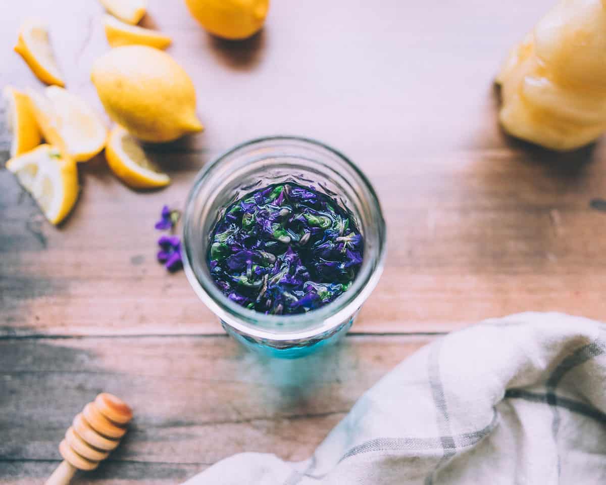 Top view of violet flowers steeping in water that is turning bright blue, on a wood surface surrounded by lemons, a honey bear, and a honey stick. 