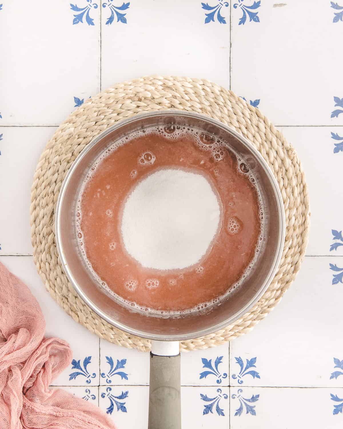 A pan of pink rhubarb syrup with white sugar in the middle ready to cook and dissolve. On a woven trivet on a white surface with blue designs. 