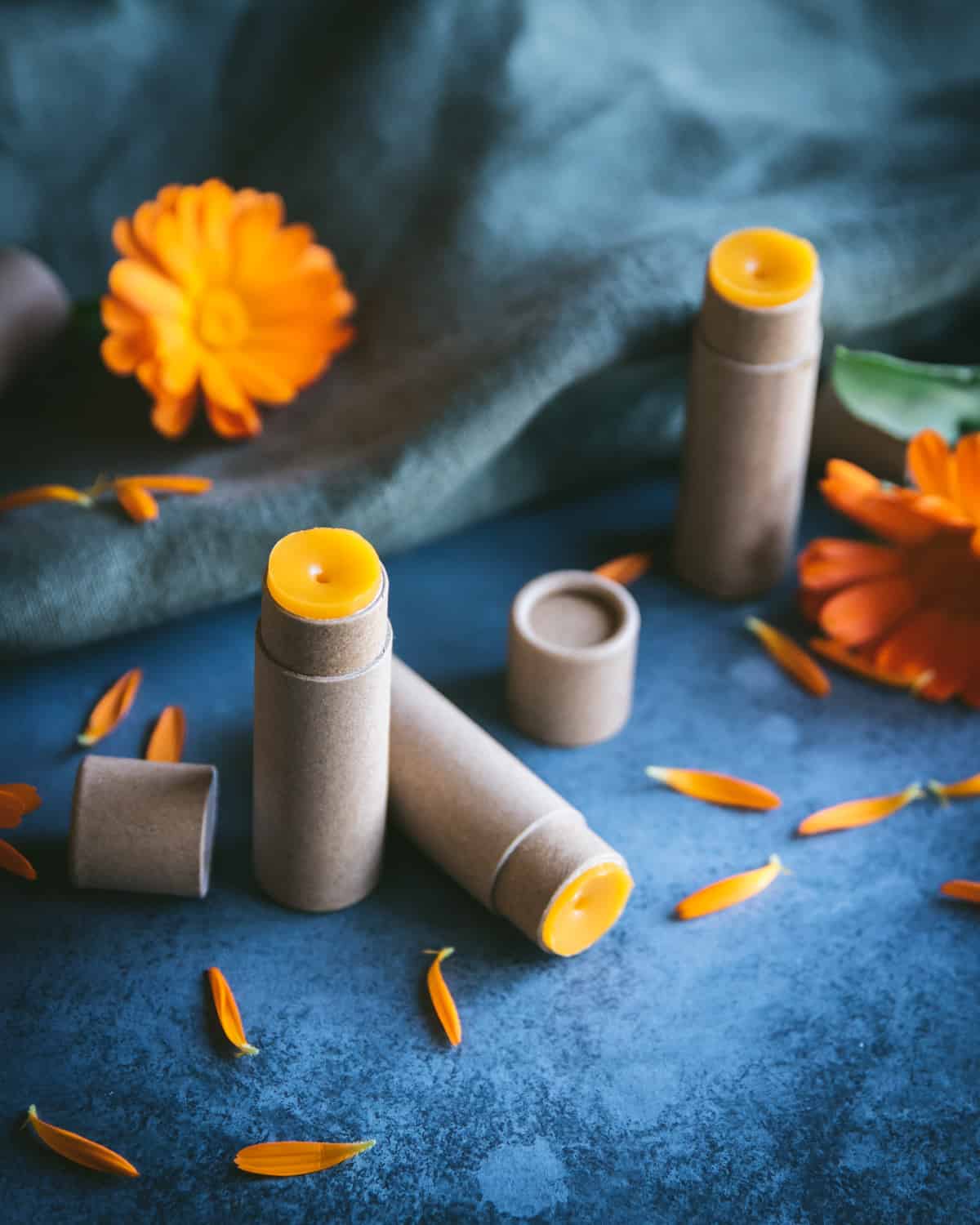 2 Calendula lip balms in paper tubes, on a dark blue cloth surrounded by calendula flowers and petals. 