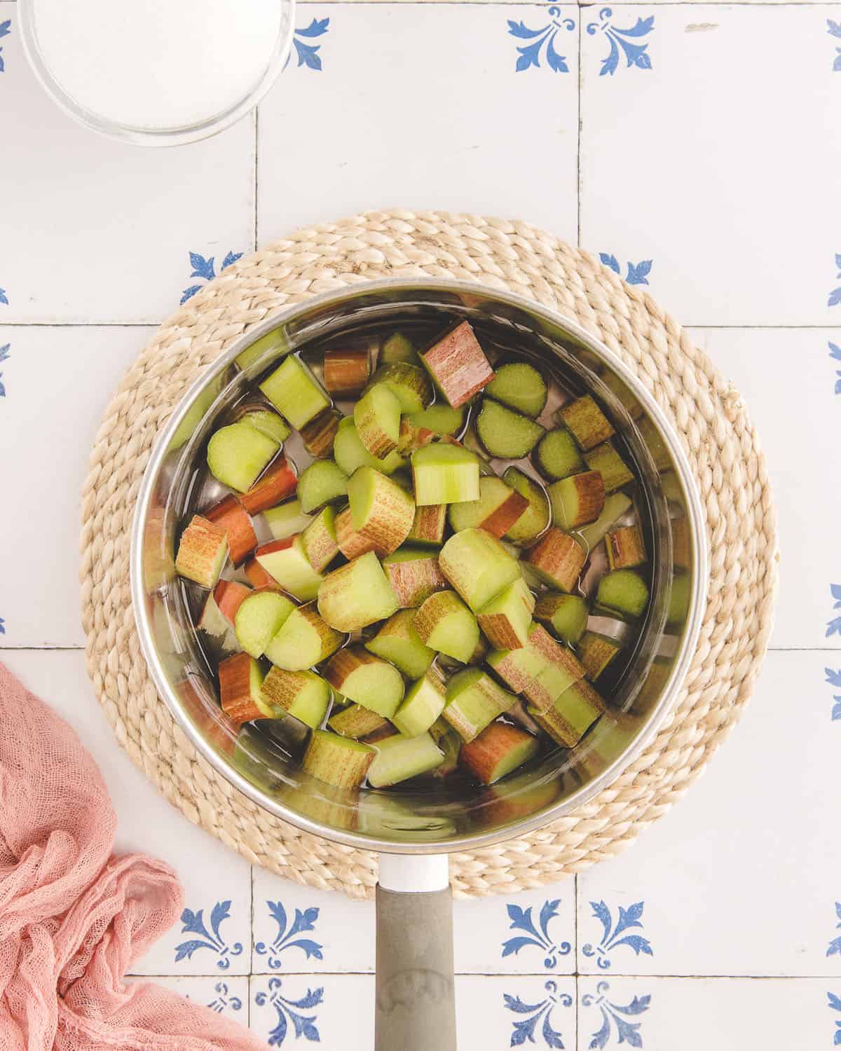 Rhubarb chopped in a pan sitting on a woven circle trivet, on a white surface with blue designs. 