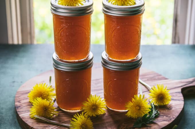 4 jars of sealed dandelion jelly, stacked in twos. On a circular wood cutting board surrounded and topped by fresh dandelion flowers, in front of a window with natural light.