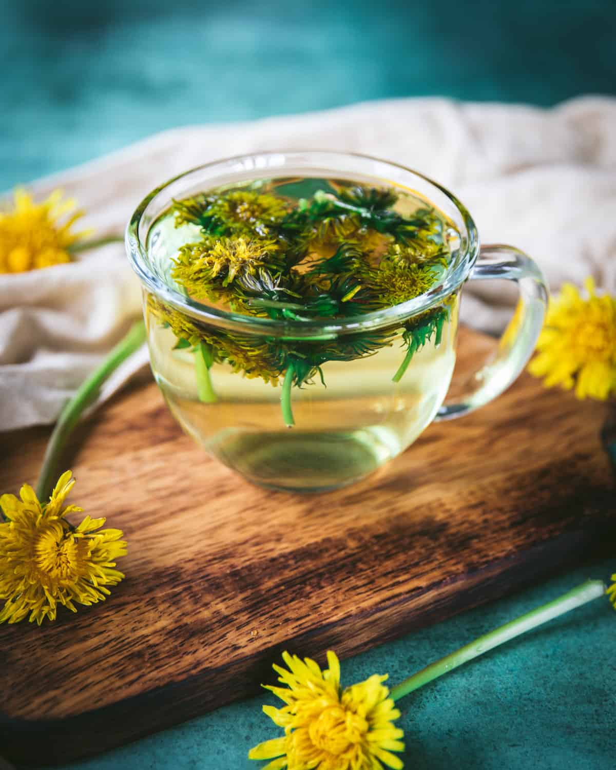 A clear mug of dandelion tea with the flowers floating on the top, on a wood cutting board with a turquoise background and surrounded by fresh dandelion flowers. 