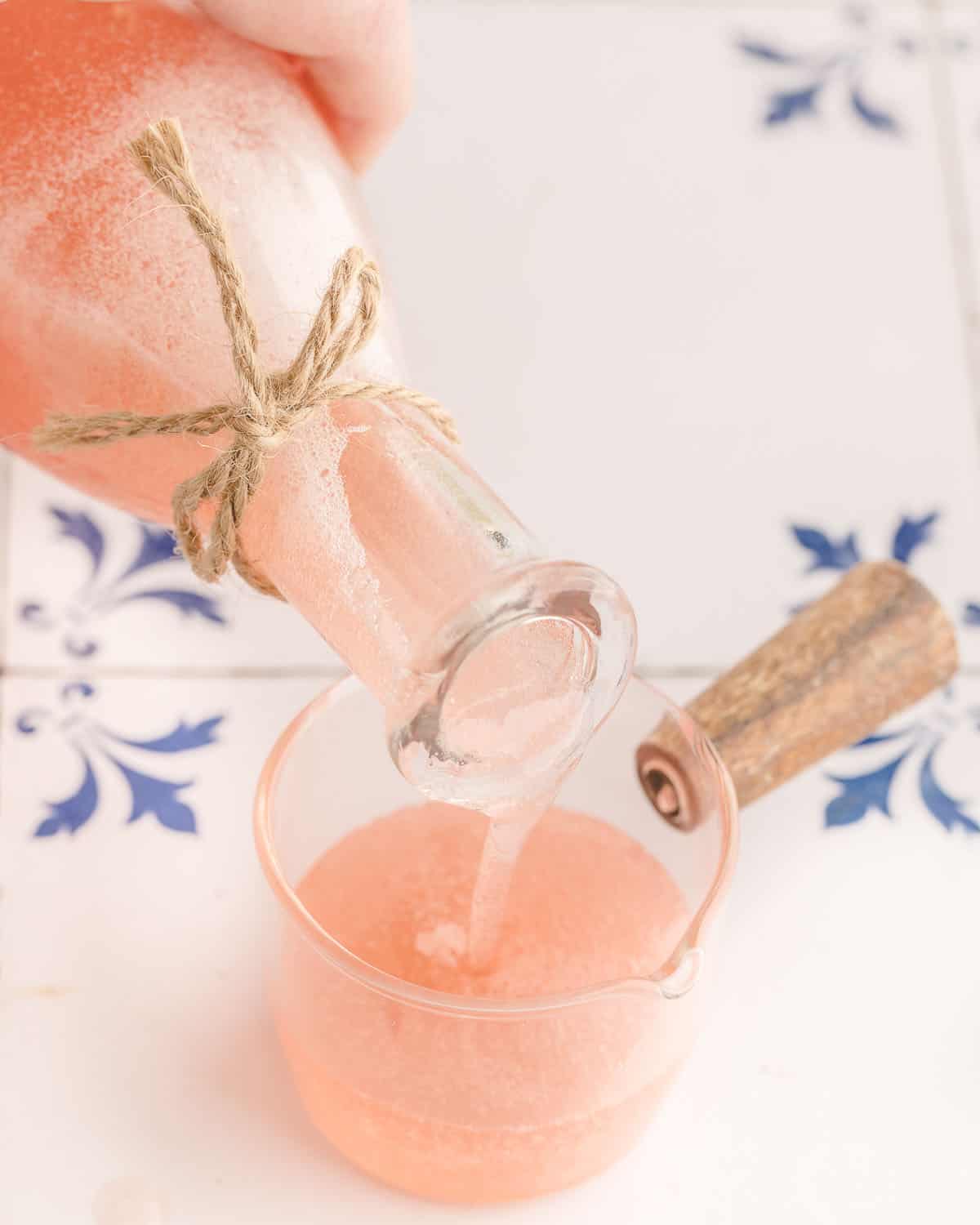Pink rhubarb syrup pouring into a glass along with a cocktail mix, on a white surface with blue designs. 
