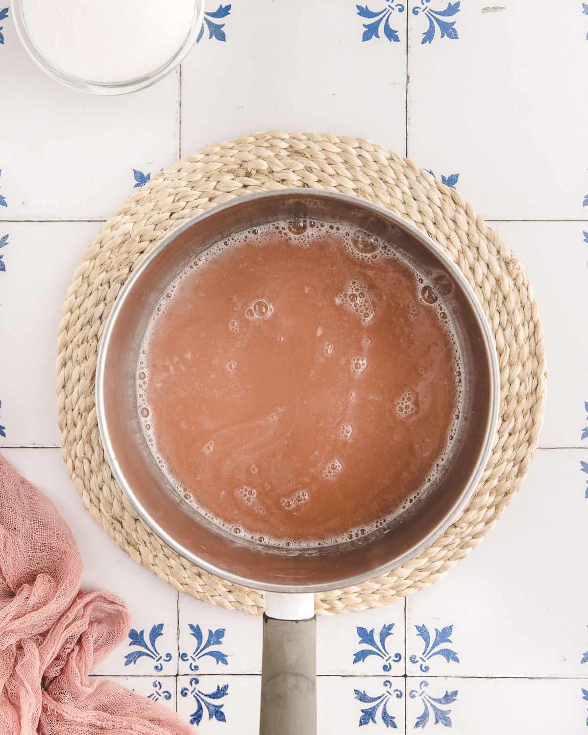 Pink rhubarb liquid in a pan with simmering bubbles. On a white surface with blue designs. 