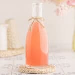 A bottle of pink rhubarb syrup with a bow of twine tied onto the bottleneck, on a woven doily with a white and light background.