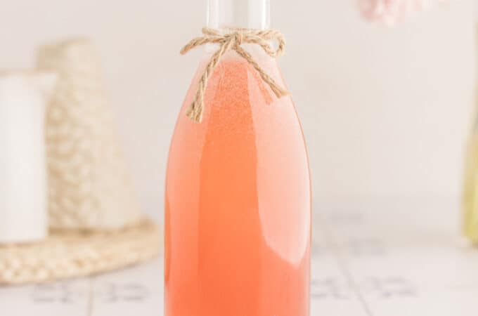 A bottle of pink rhubarb syrup with a bow of twine tied onto the bottleneck, on a woven doily with a white and light background.