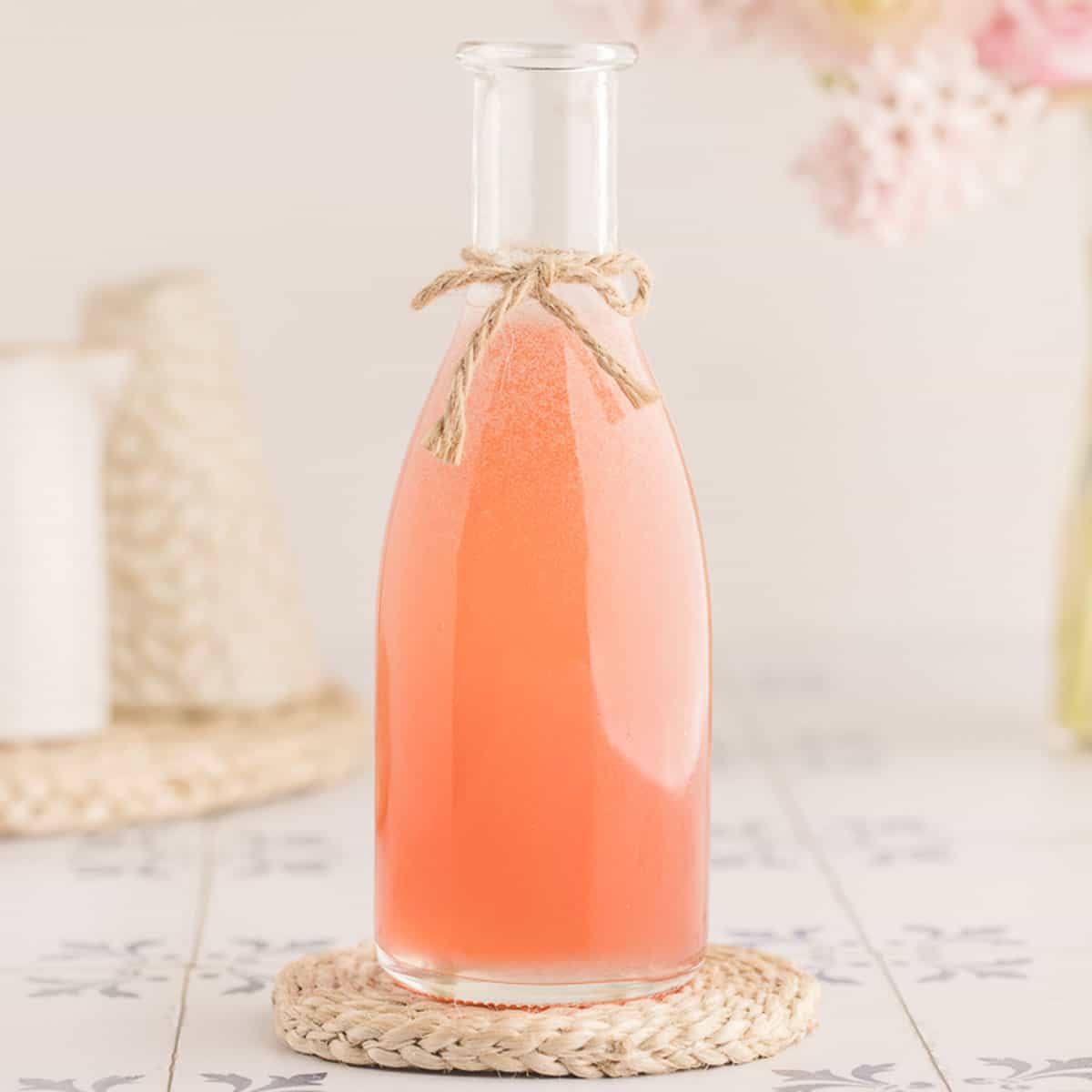 A bottle of pink rhubarb syrup with a bow of twine tied onto the bottleneck, on a woven doily with a white and light background. 