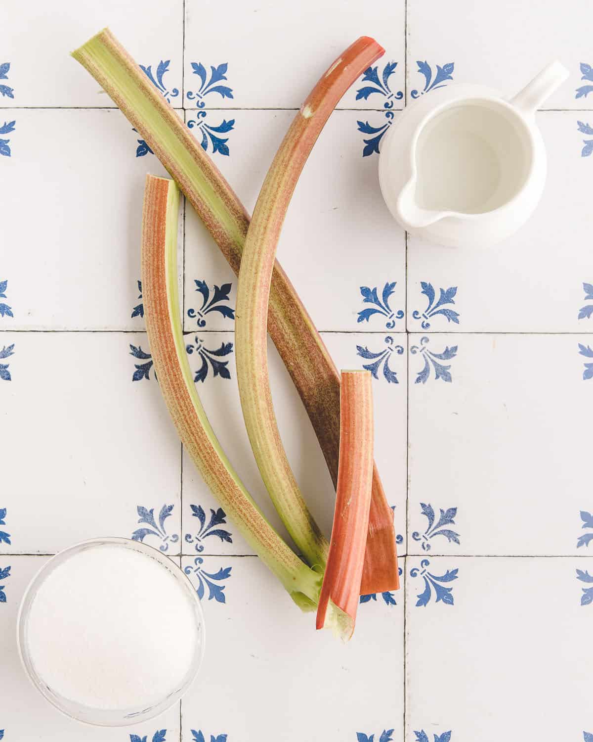 Fresh pink and green rhubarb stalks with a sugar bowl beside it, on a white background with a blue design. 