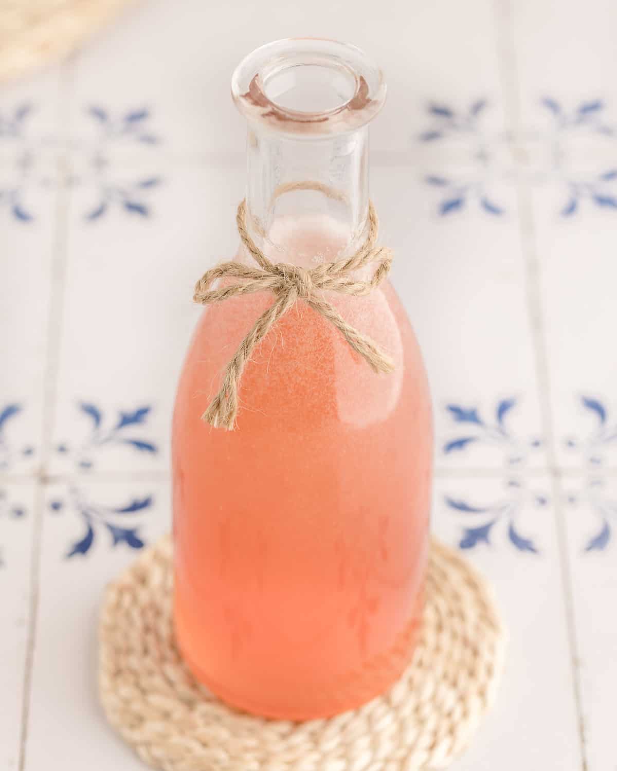 Rhubarb syrup that is pink in a glass bottle with a twine bow tied to it, on a small woven trivet on a white counter surface with blue designs on it. 