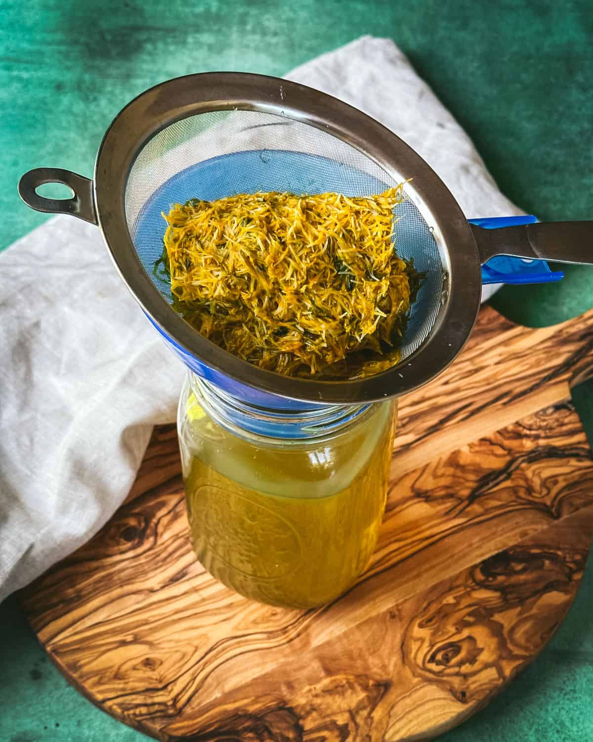 A mesh strainer with dandelion petals over a jar with dandelion tea on a wood cutting board with a natural cloth and green background.