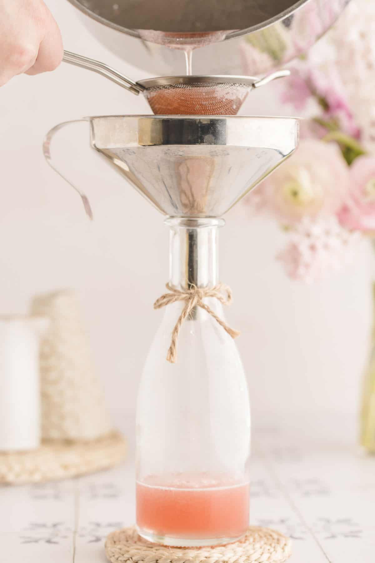 Pink rhubarb syrup pouring through a silver funnel into a bottle with a twine bow on it. Light and bright with a white background and light pink flowers in the background.