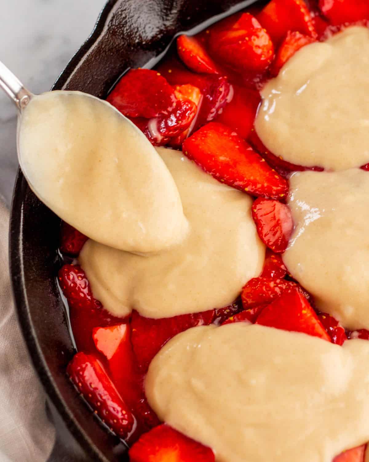 Close up of sugared strawberries with cobbler batter being poured in dollops on top in a cast iron skillet. 