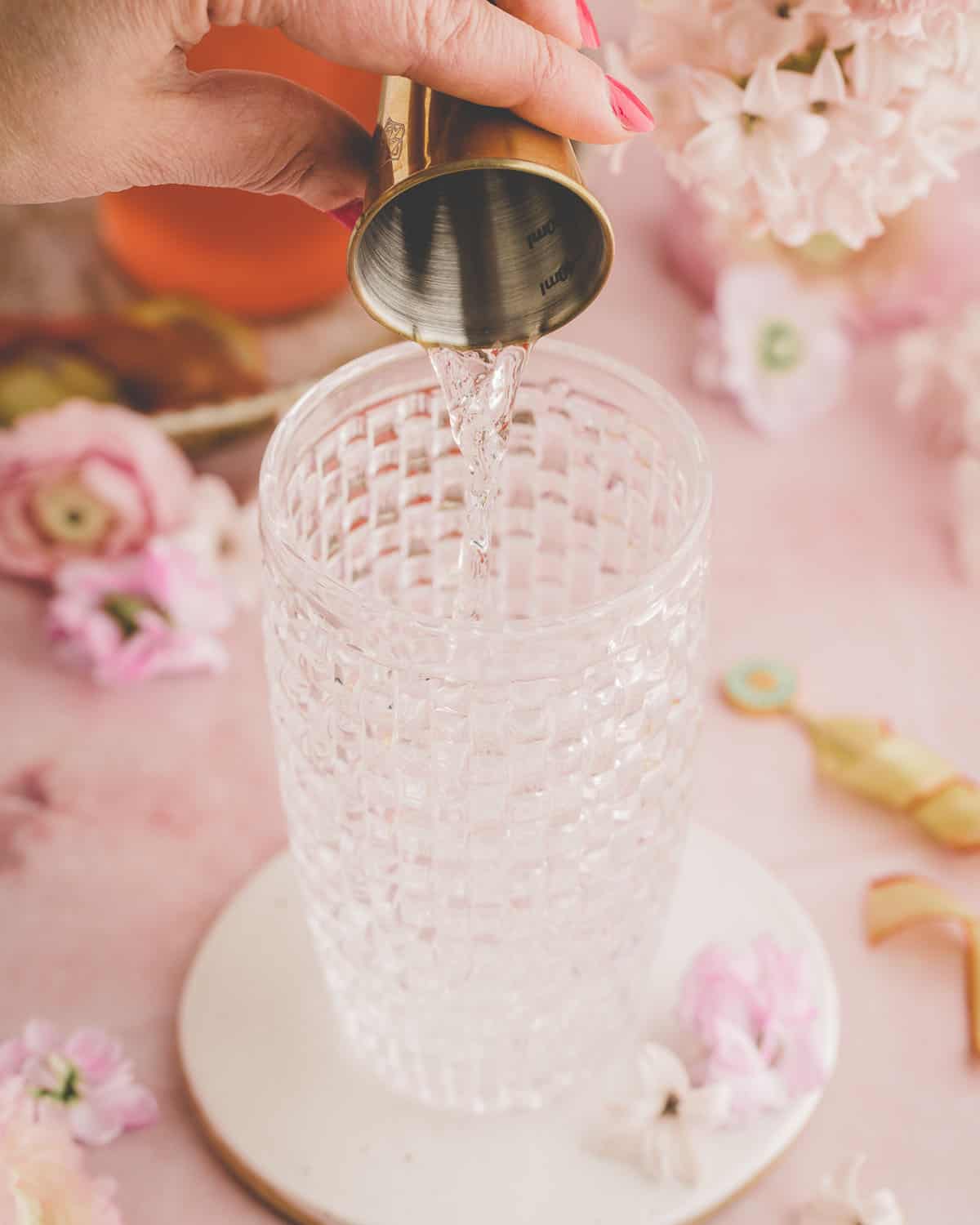 A shot of gin pouring into a vintage glass shaker, with a pink background surrounded by pink flowers and rhubarb syrup. 