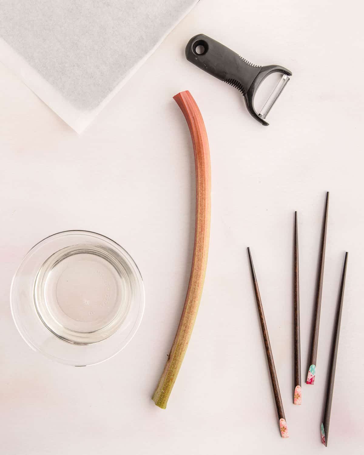 One rhubarb stalk, a peeler, and chopsticks on a light pink surface. 