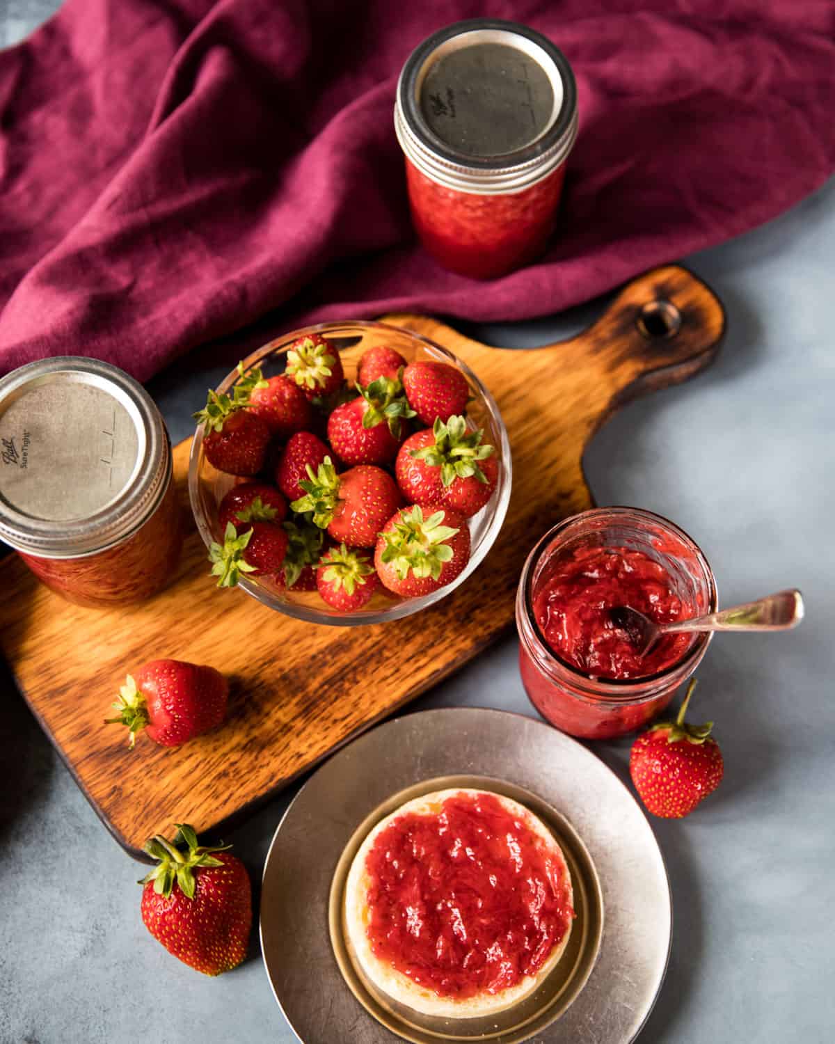 A bowl of fresh strawberries, a jar of opened strawberry jam with a spoon on a wooden cutting board. Surrounded by burgundy fabric and a plate with jam covered toast. 