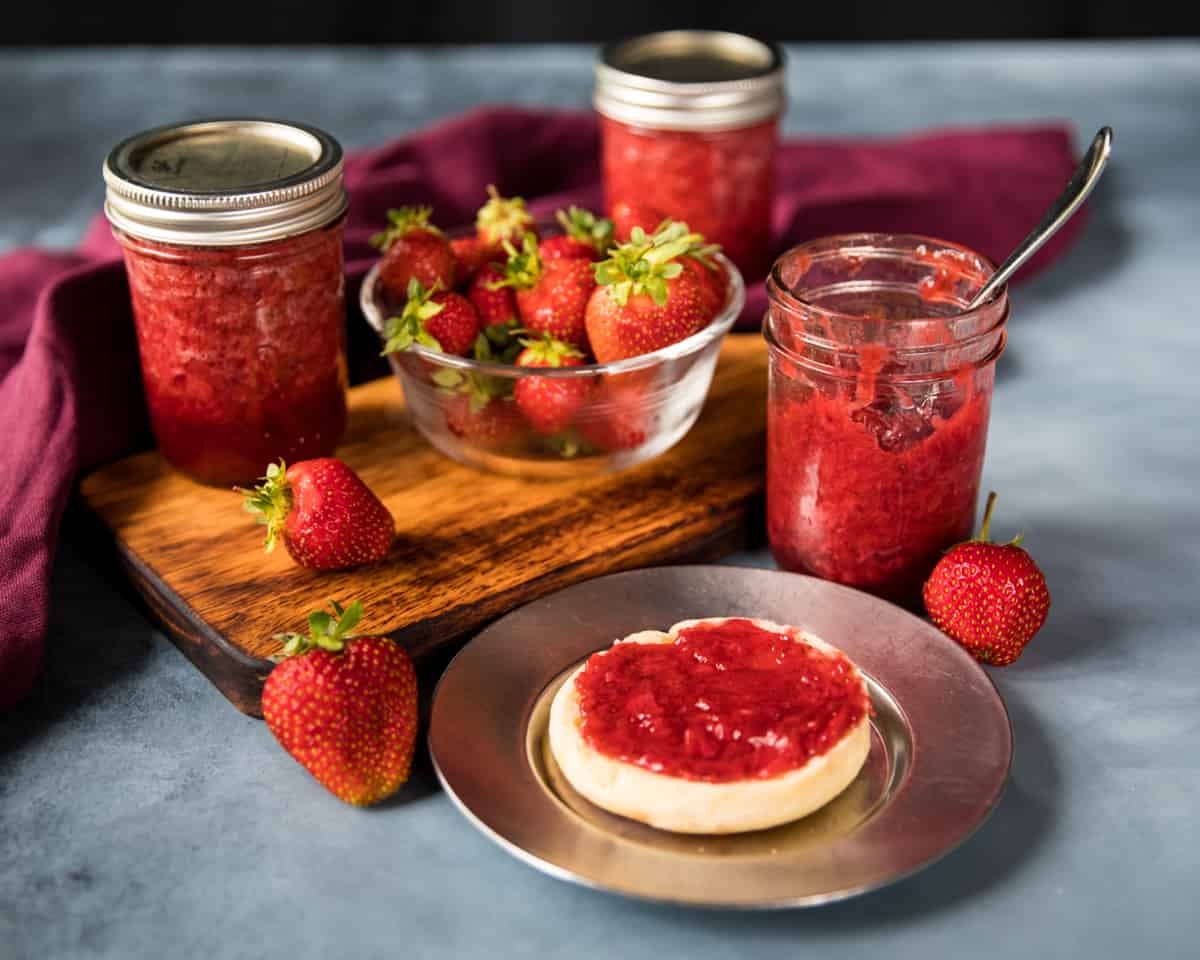A plate with an english muffin with strawberry jam on it, surrounded by jars of jam and fresh strawberries. 