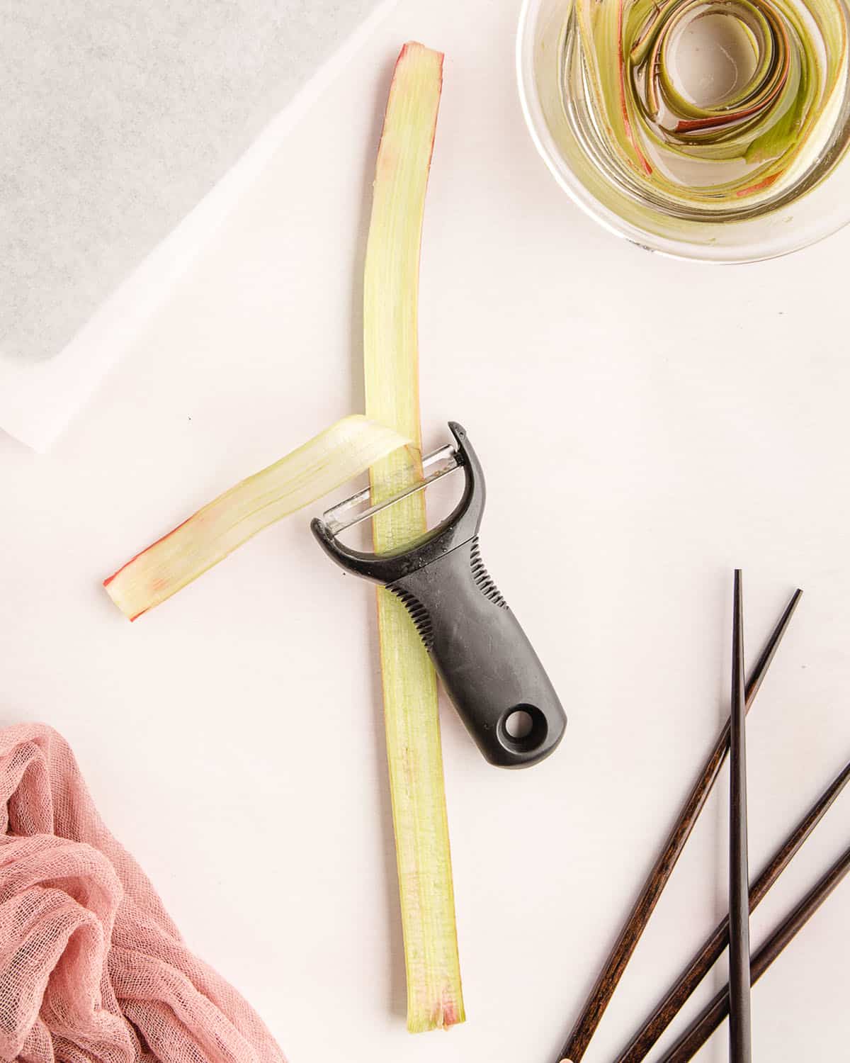 A peeler in process of peeling rhubarb into a thin flat strip, surrounded by pink fabric, chopsticks, and a bowl of simple syrup. 