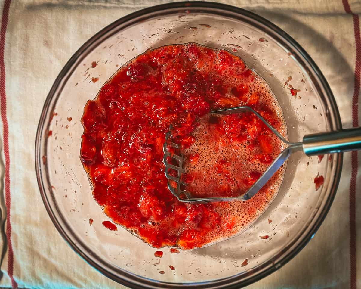 A potato masher mashing up the strawberries in a bowl. 