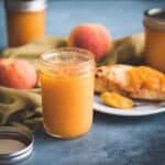 A jar of peach jam surrounded by a plate of toast with peach jam, whole peaches, and more jars of peach jam. On a blue surface with a natural colored cloth weaving through.