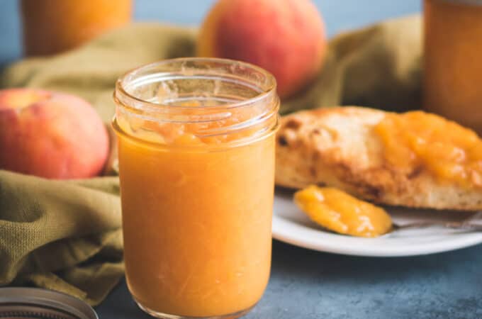 A jar of peach jam surrounded by a plate of toast with peach jam, whole peaches, and more jars of peach jam. On a blue surface with a natural colored cloth weaving through.