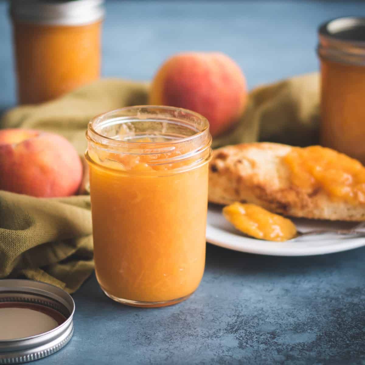 A jar of peach jam surrounded by a plate of toast with peach jam, whole peaches, and more jars of peach jam. On a blue surface with a natural colored cloth weaving through.