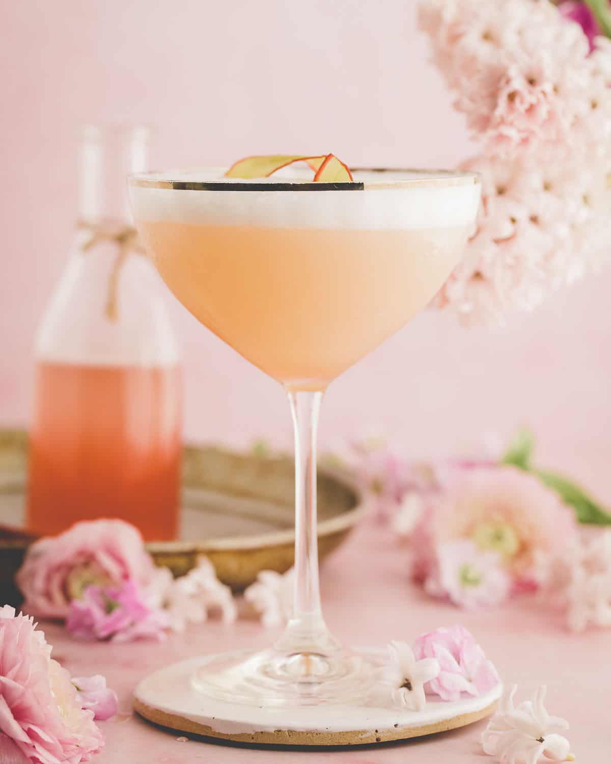 A vintage champagne glass with a silver rim filled with foamy rhubarb gin sour, on a pink surface surrounded by flowers and a bottle of rhubarb syrup. 