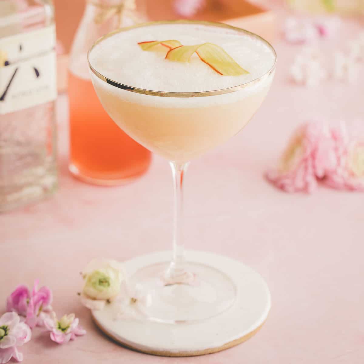 A cocktail glass with a silver rim, containing a pink rhubarb cocktail drink, garnished with a candied rhubarb ribbon. On a pink surface, surrounded by pink flowers and a bottle of rhubarb syrup. 