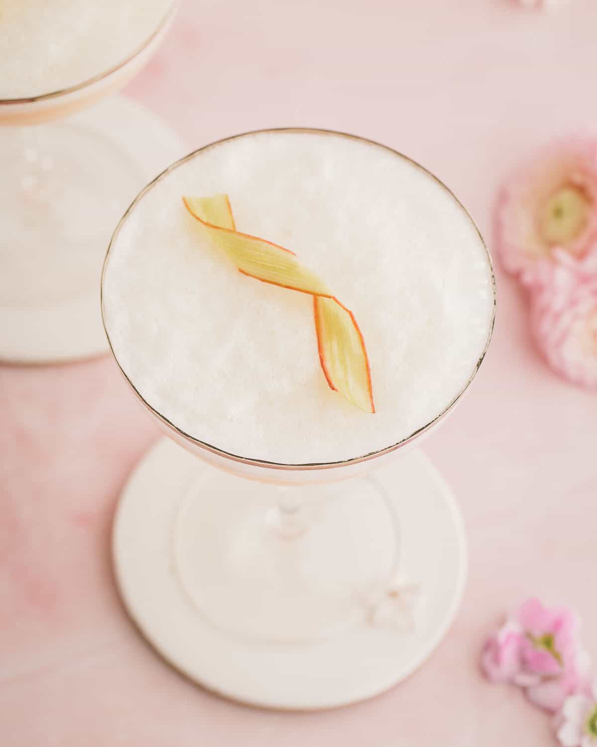 Top view of rhubarb gin sour with foam and candied rhubarb ribbon garnished, on a pink surface surrounded by pink flowers. 