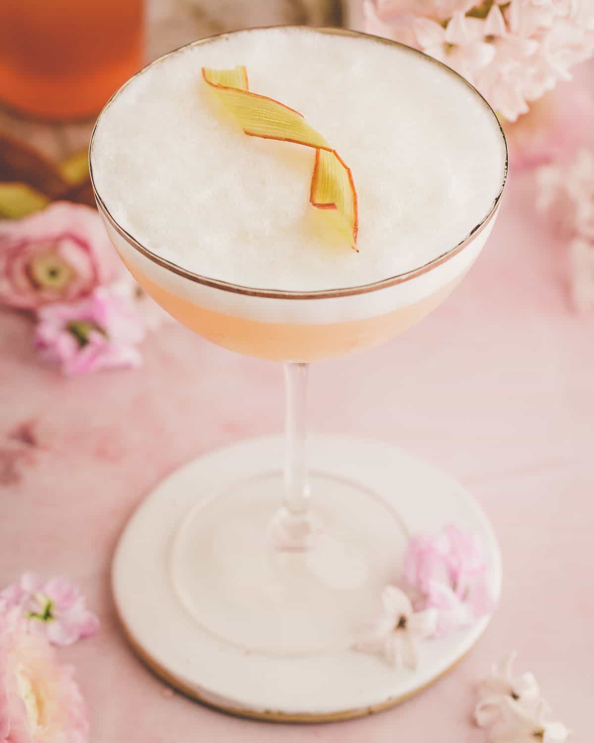 A rhubarb gin sour cocktail with a foamy top and a candied rhubarb ribbon to garnish, on a pink surface with pink flowers surrounding. 