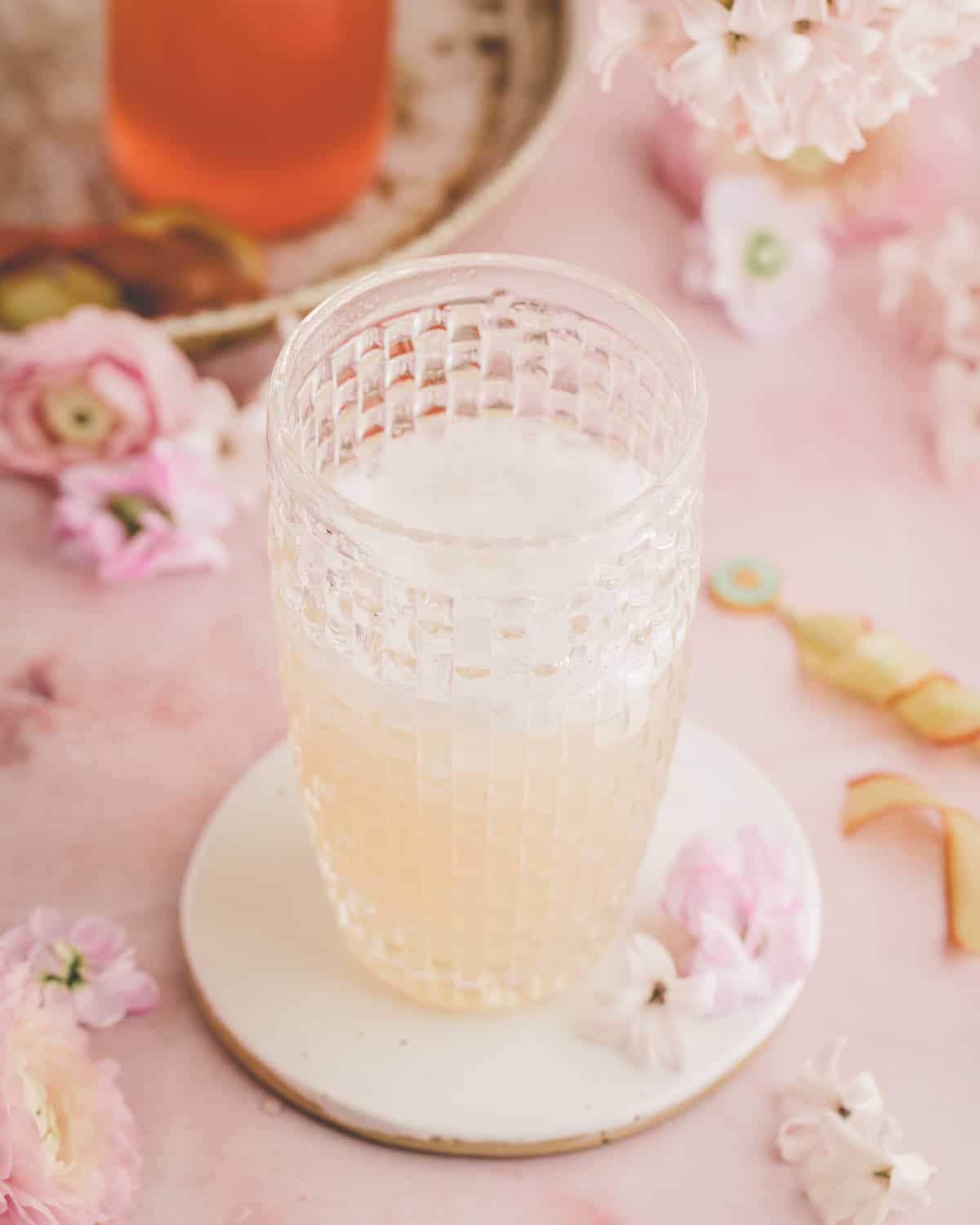 A vintage glass cocktail shaker with pink rhubarb cocktail in it that has a layer of foam on top, on a pink surface surrounded by pink flowers. 
