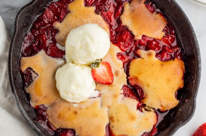 Strawberry cobbler in a cast iron skillet, on a white surface surrounded by fresh strawberries.
