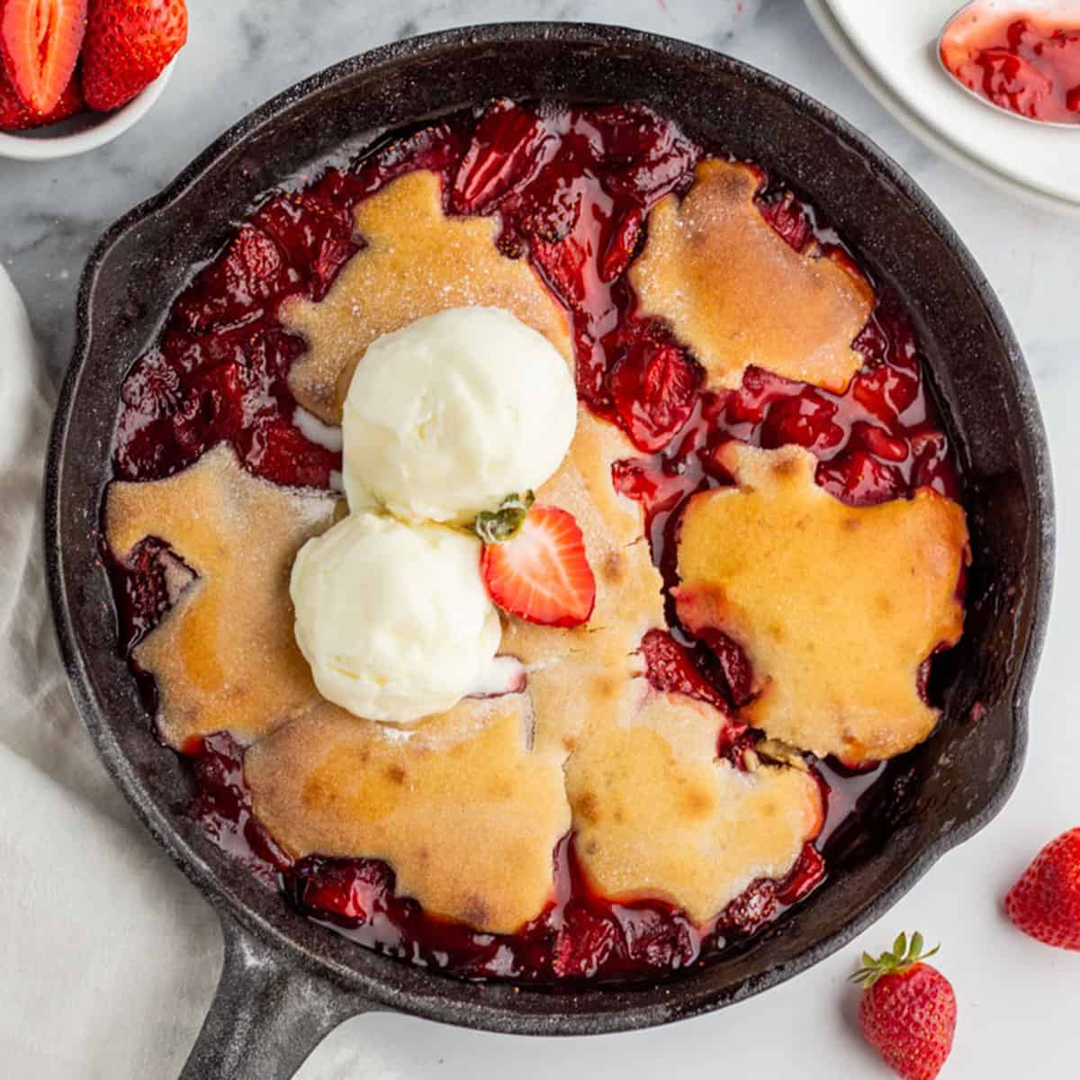 Strawberry cobbler in a cast iron skillet, on a white surface surrounded by fresh strawberries. 