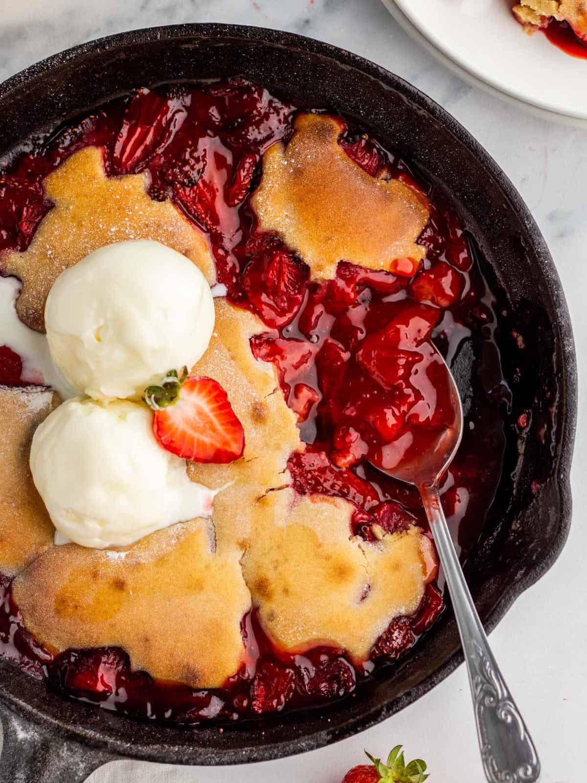 Strawberry cobbler in a skillet topped with vanilla ice cream and a fresh strawberry, with a spoon dipping into the cobbler. 