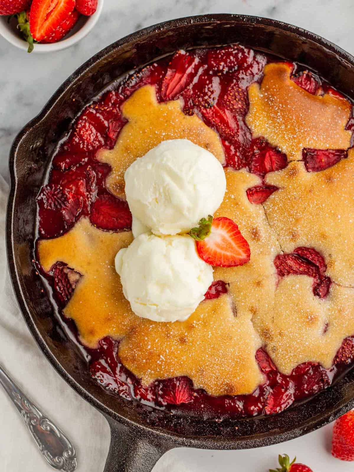 Strawberry cobbler in a skillet with 2 scoops of vanilla ice cream and a strawberry slice on top, surrounded by fresh strawberries. 